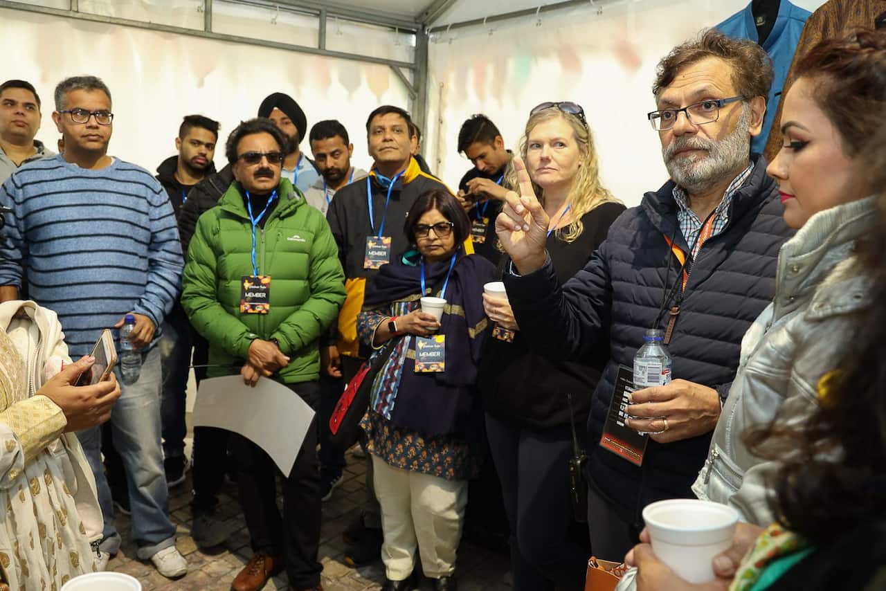 A man wearing glasses and a black puffer jacket gestures with one hand while holding a bottle of water in the other as he addresses a small group of people standing in a makeshift tent.