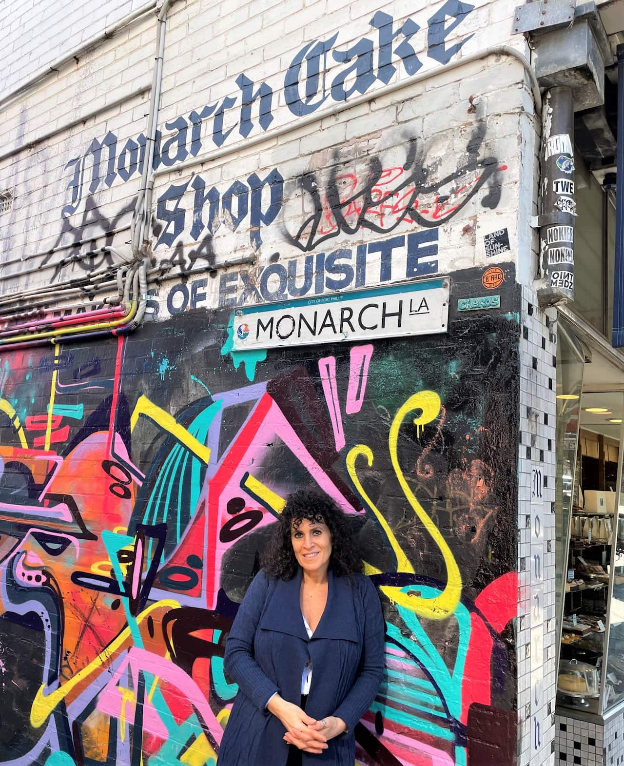 A woman in a blue cardigan stands at a street corner, against a background of graffiti.