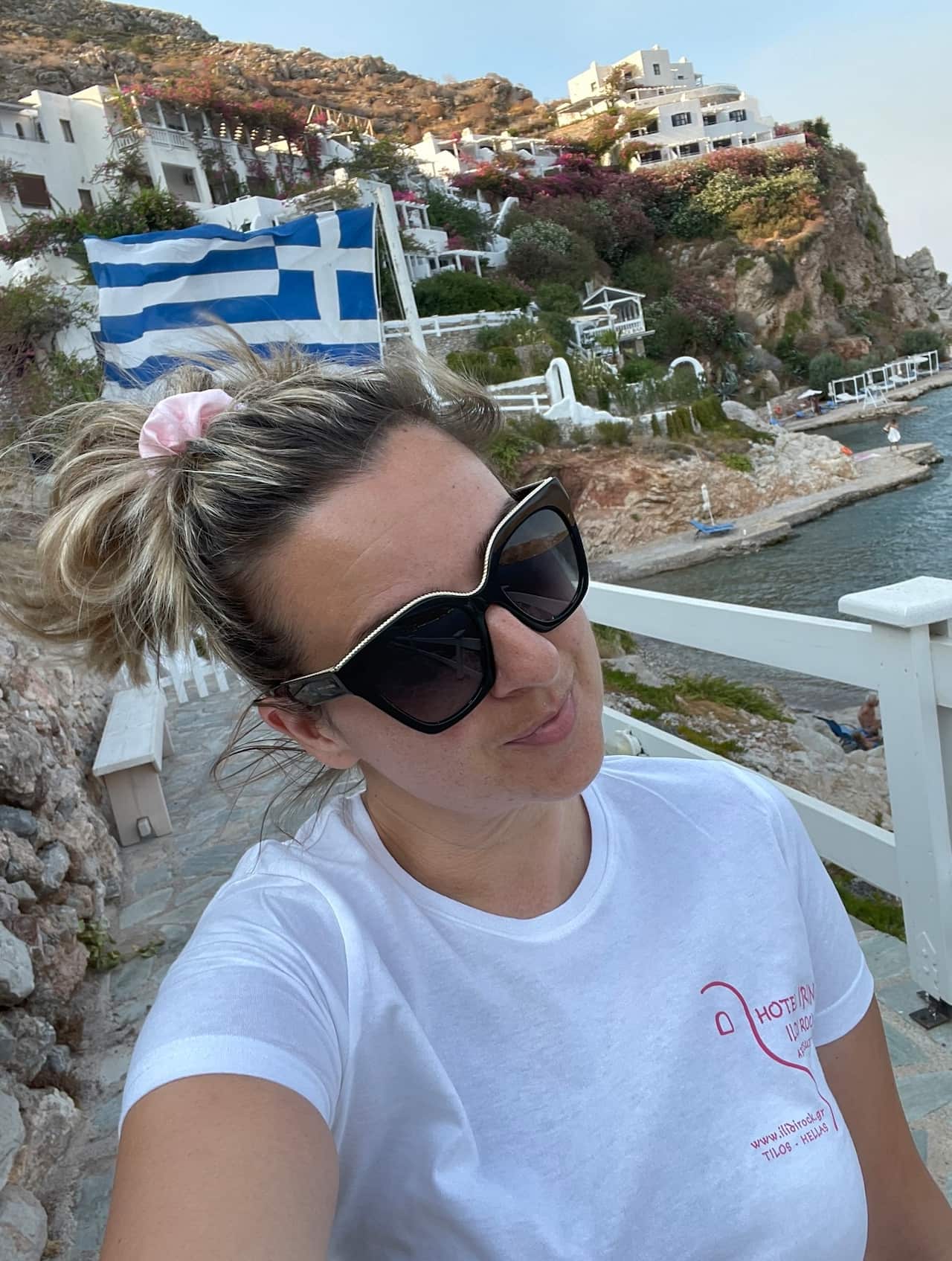 A woman in a white t-shirt wearing sunglasses taking a selfie. There's water and a cliff in the background.