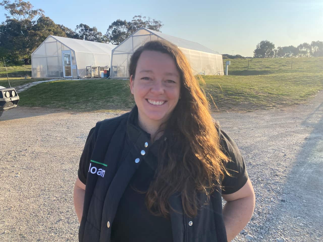 A woman wearing a vest smiles as she stands outside on a dirt road. 