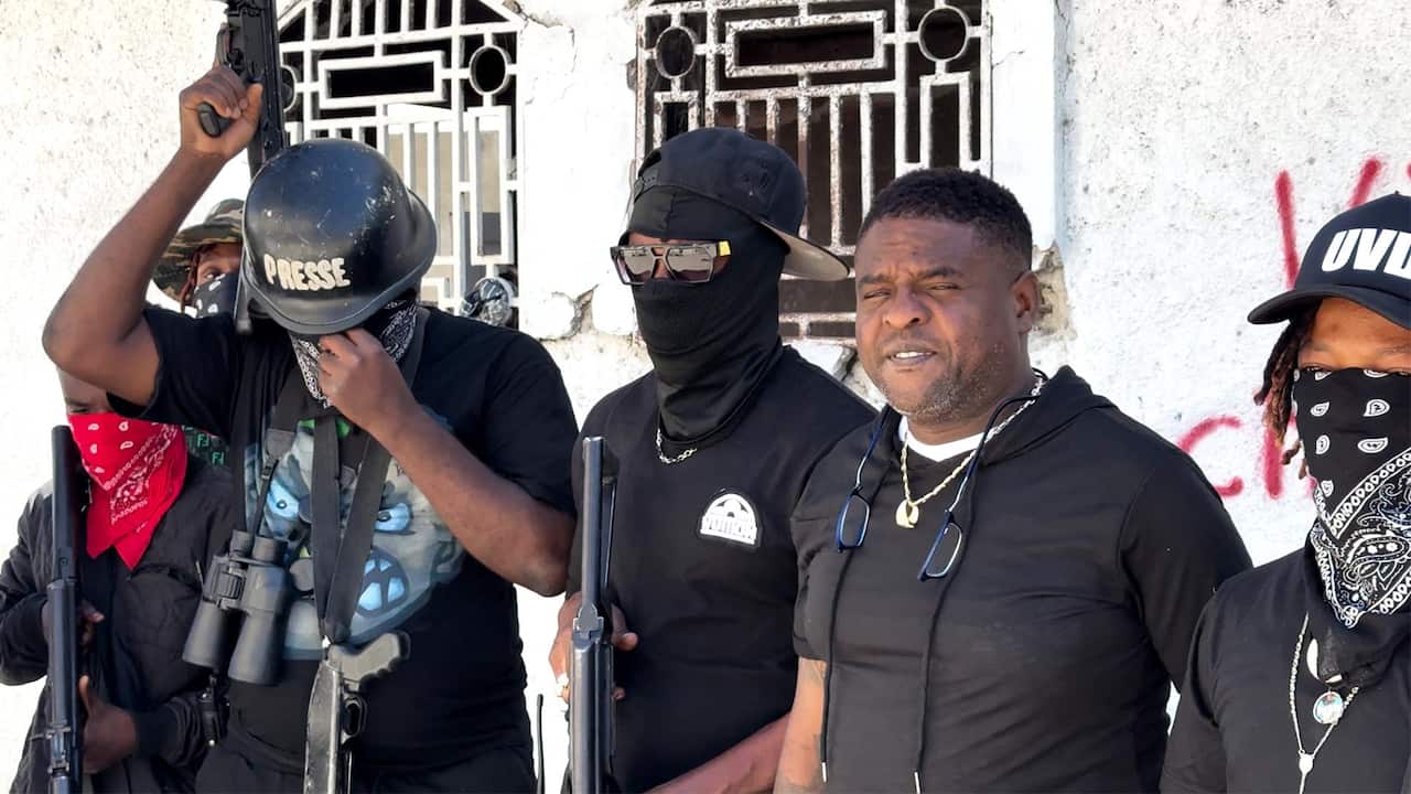 A group of men in black clothing. All but one are armed, wearing hats or baseball caps, and with their faces covered by bandanas