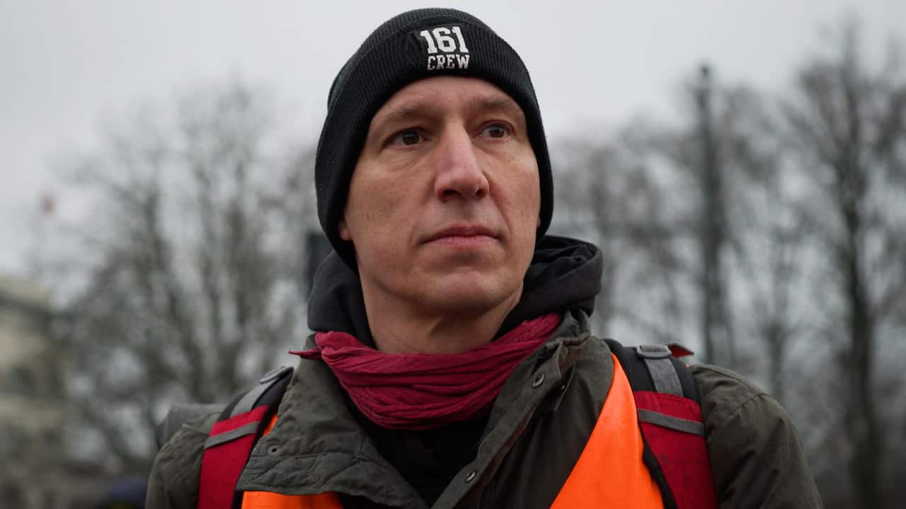 A middle-aged man wearing a dark winter jacket, red scarf, black beanie hat, orange safety vest and a backpack