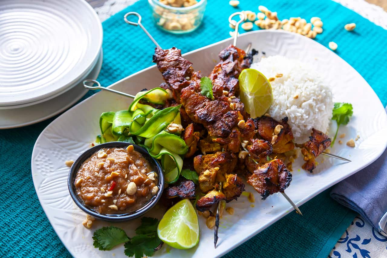 A rectangularish white platter sits on a teal coloured textured cloth. Several nicely charred chicken skewers sit on the platter, along with rice, cucumber ribbons and a shallow bowl of brown sauce. 