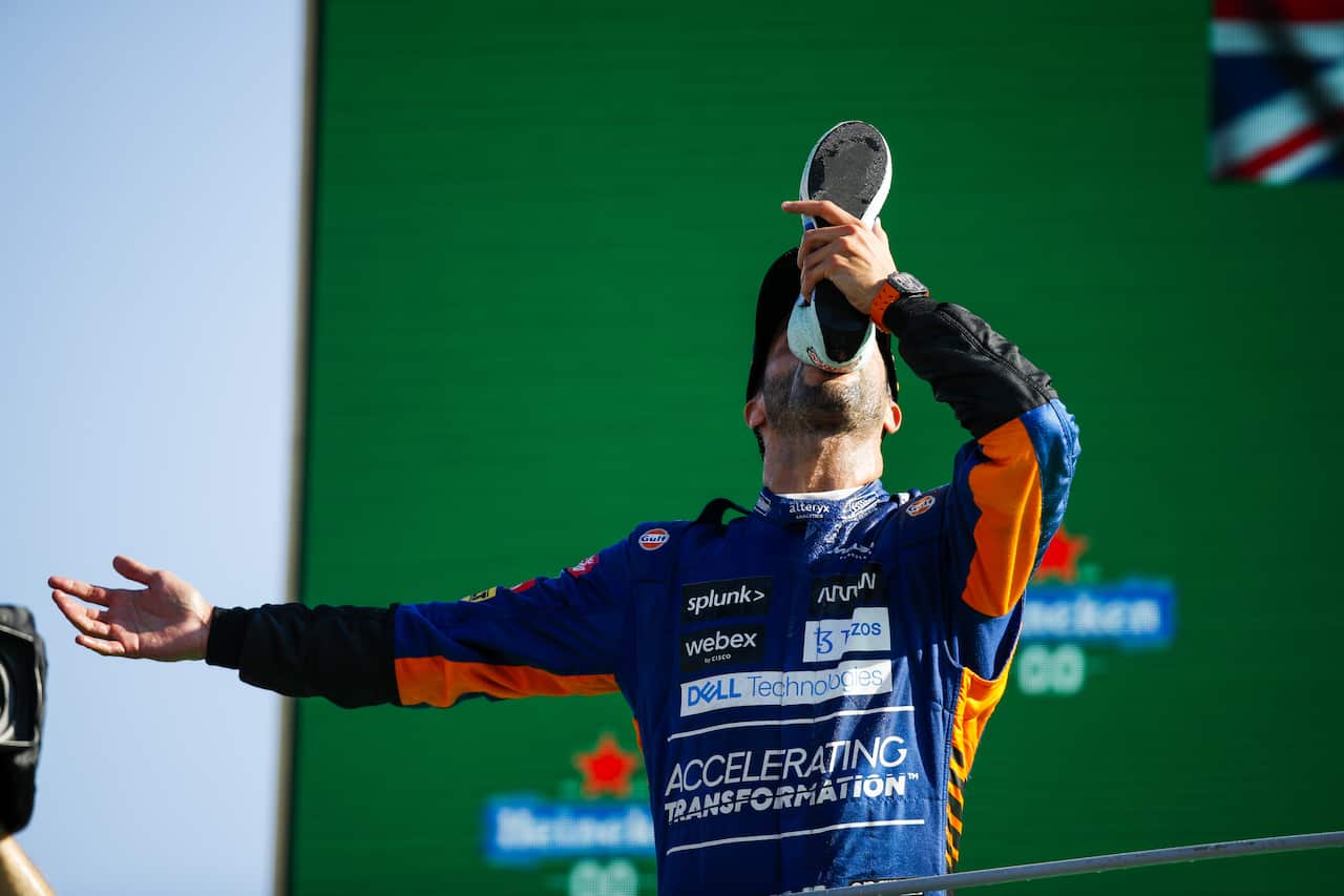 A man in a race car driver jumpsuit drinks beer out of a shoe.