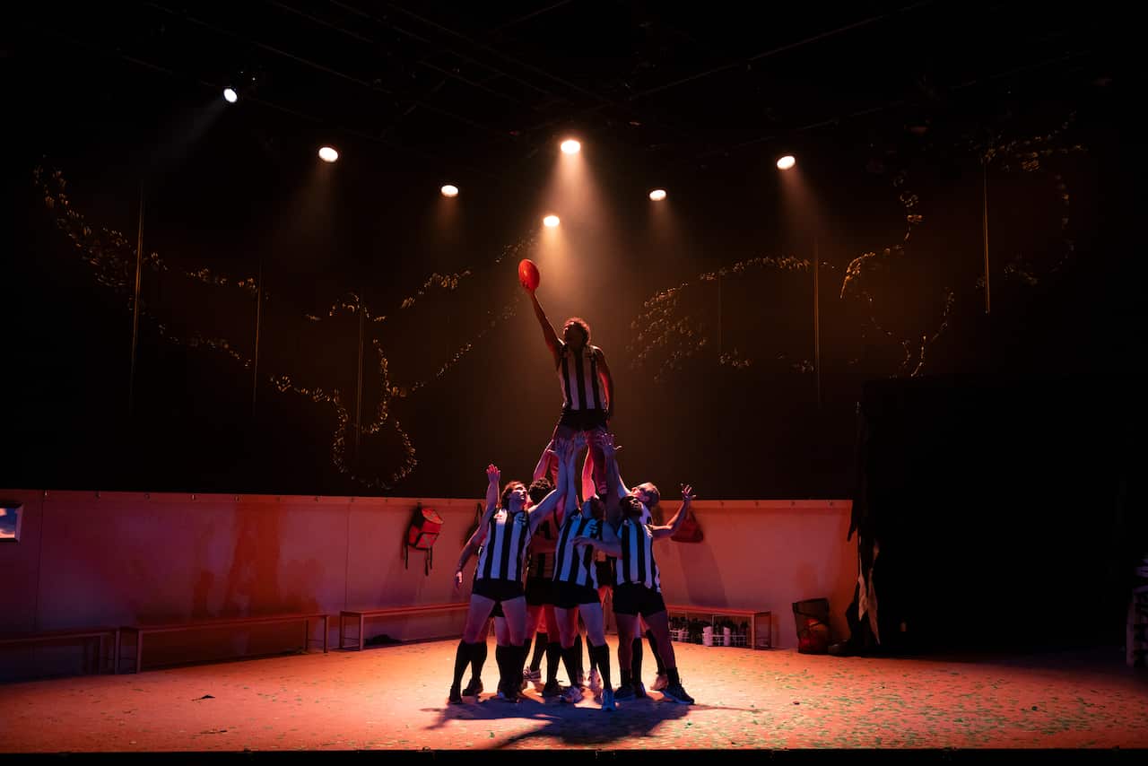 A cast of actors on stage, one man being held up by the others to grab a football.