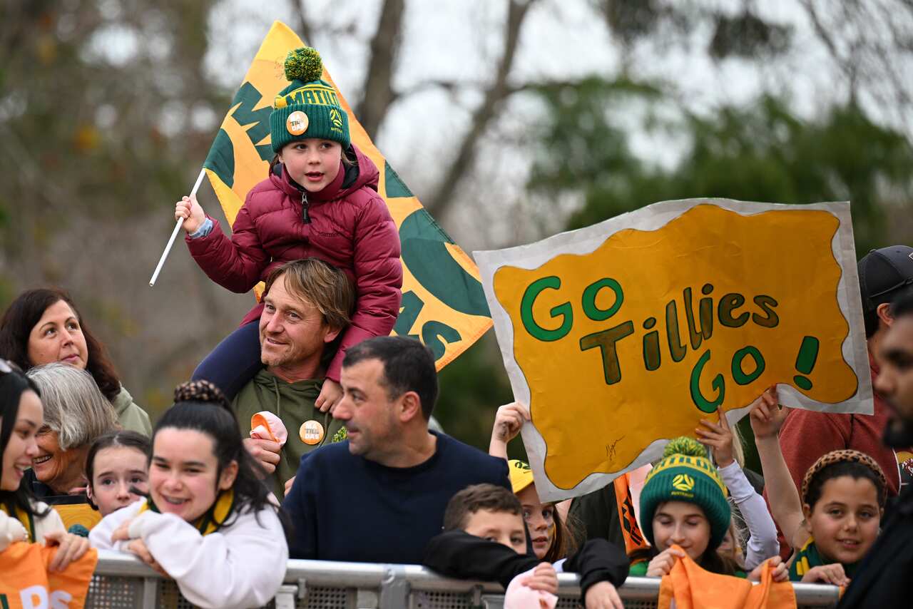 A group of people cheering for the Matildas, including a man carrying a child on his shoulders, with flags and a banner that reads "Go Tillies go!". 