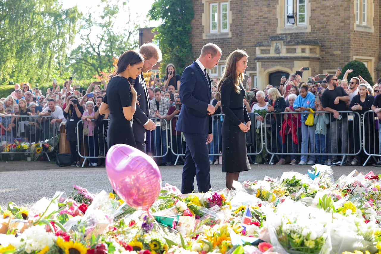 Two men and women wearing black look down at bunches of flowers arranged on the ground.