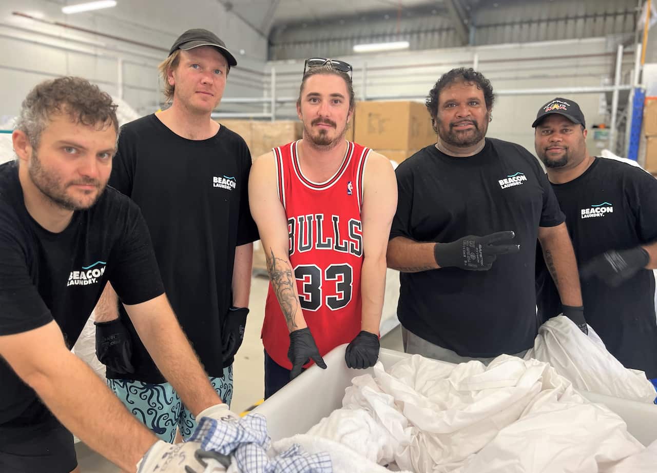 A group of men in t-shirts stand around a trolley filled with white linen.
