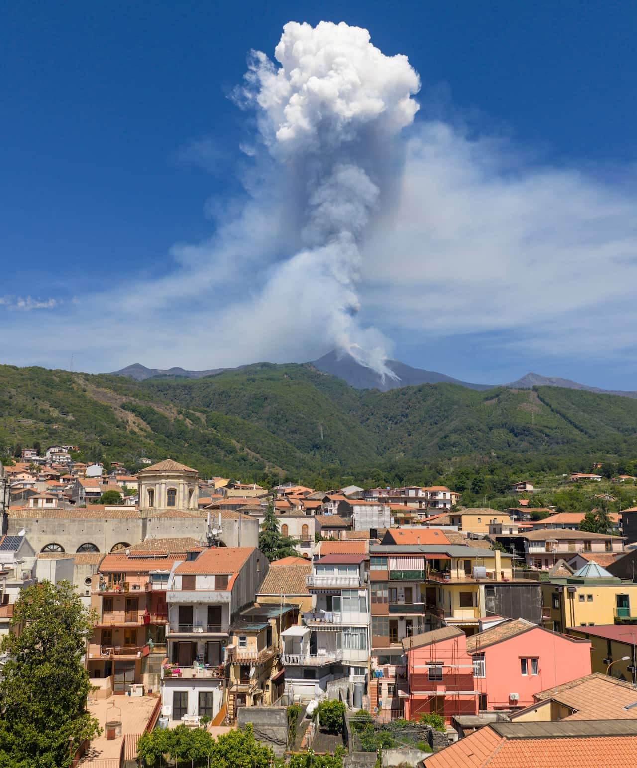 Smoke rising from a volcano, with houses seen in the foreground.