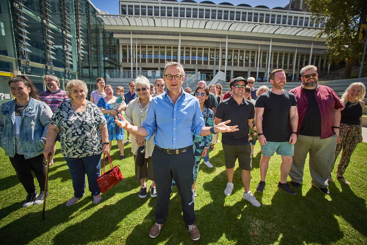 A smiling man in glasses and a blue shirt, stands with an open hands gesture in front of a large group of people on a grass lawn.