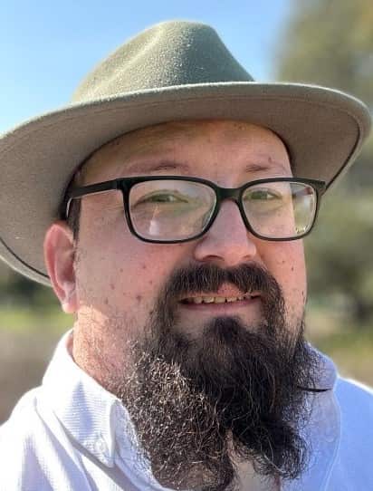 A middle-aged man with a full dark beard and glasses smiles at the camera, wearing a light-coloured collared shirt and a muted olive-green fedora. The outdoor photo is taken on a sunny day with a bright blue sky and blurred greenery, including a large tree, in the background.