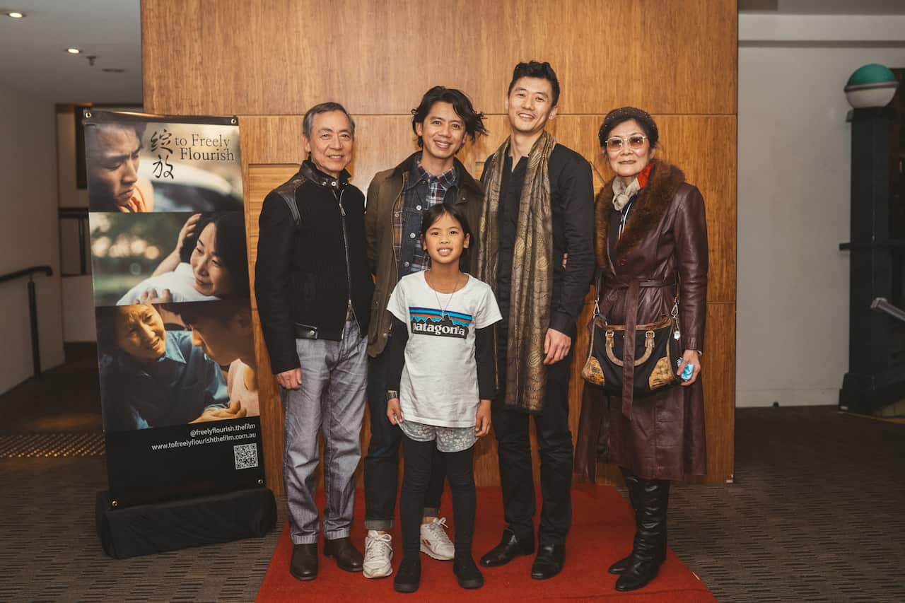 Four adults and a young child pose for a picture, standing next to a film poster and in front of a wooden background.