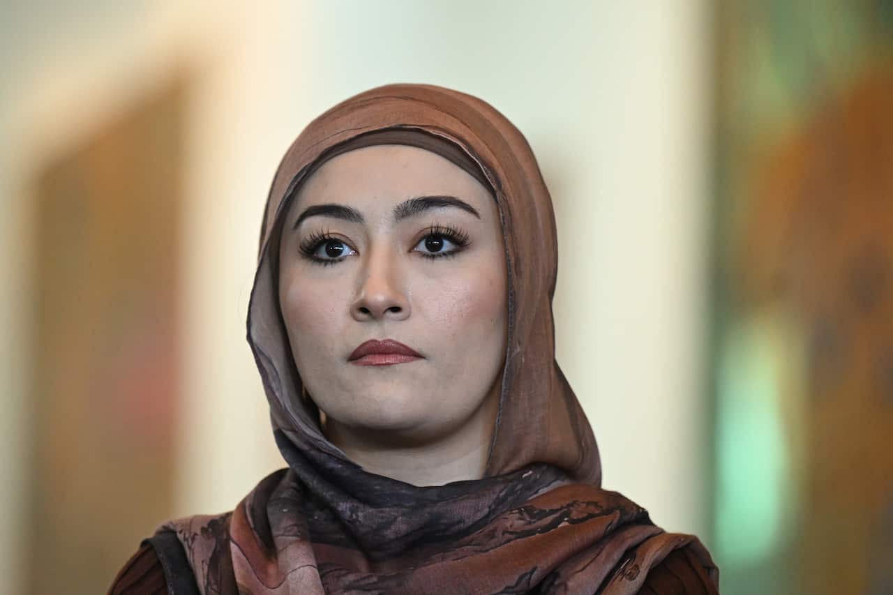 A woman wearing a brown hijab looks forward with a serious expression on her face. 