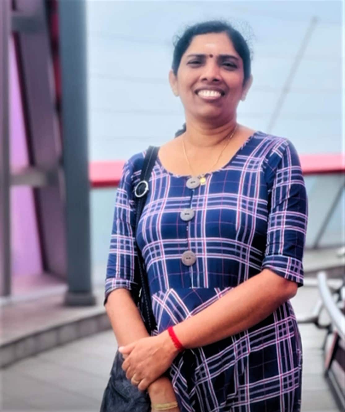 A woman in a purple checked dress stands with her hand holding her arm.