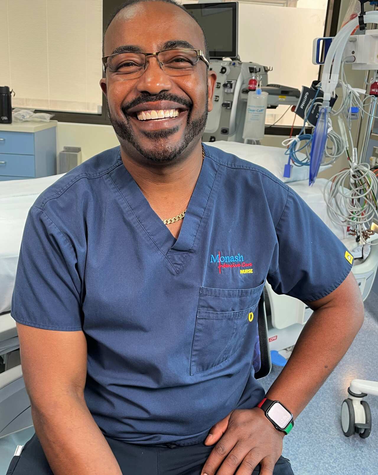 A man in a blue uniform sits in a hospital room smiling at camera.