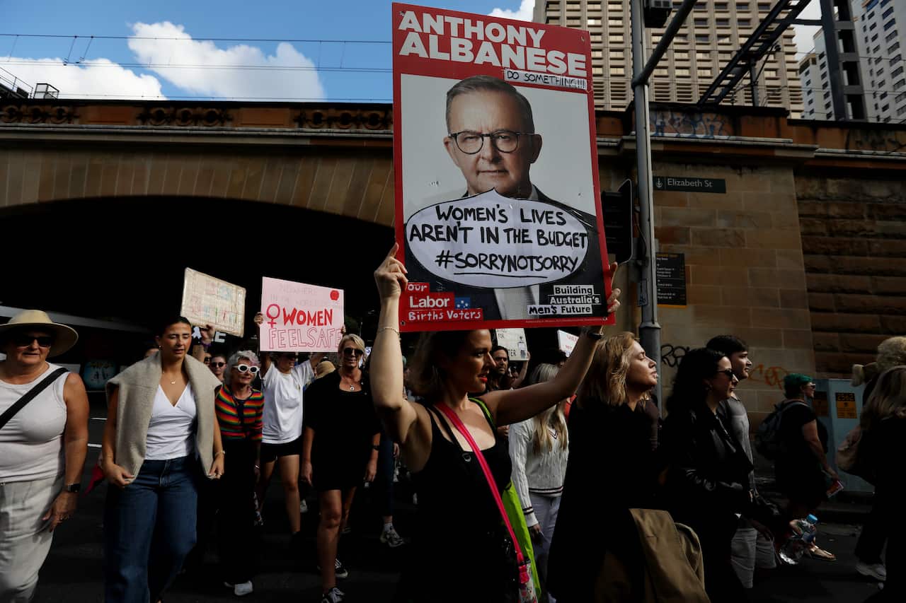 A crowd of people march at a protest, holding signs, including one with a picture of Anthony Albanese.