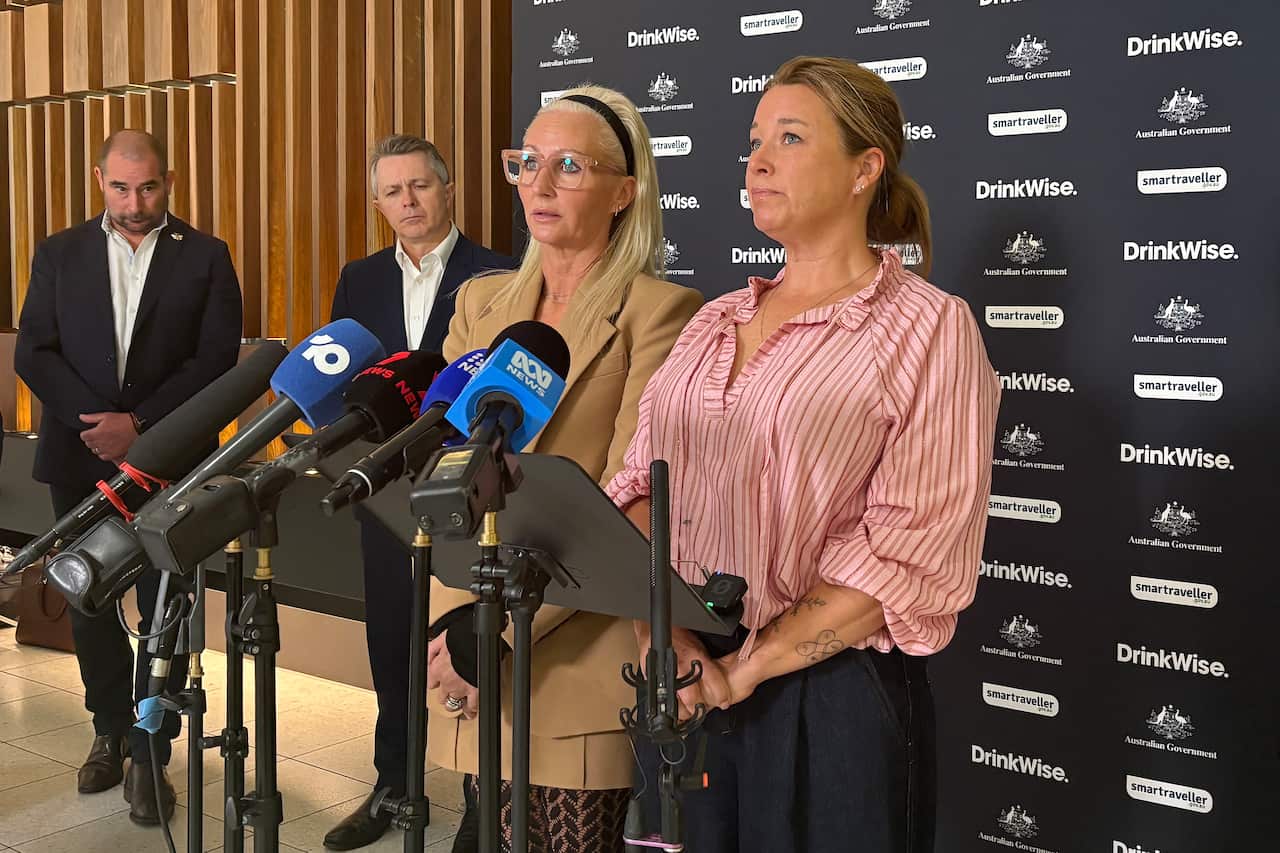 Two women stand at a lectern, with several television microphones, looking upset.