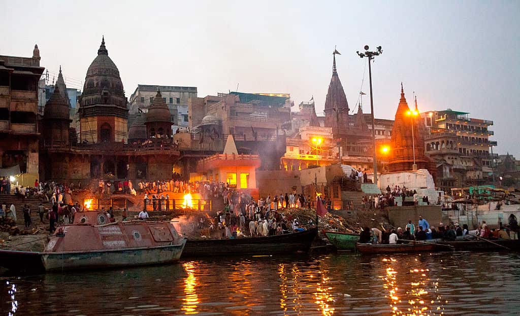 The banks of a river with buildings on it. Small fires are being burned. The water is in the foreground.