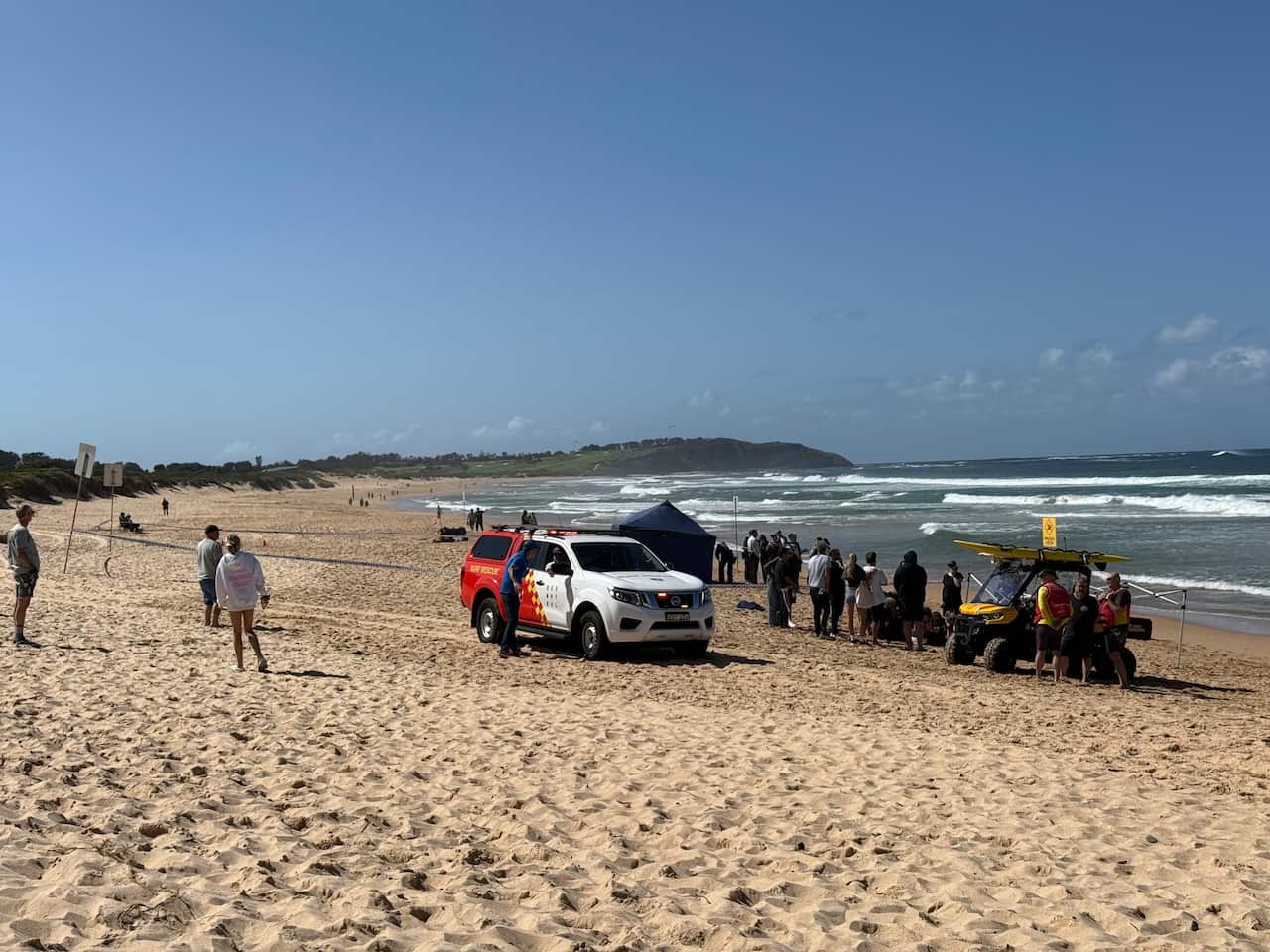 A rescue truck is parked on a sunny beach, with a crowd of people gathered around a nearby tent. In the background, waves are crashing on the shore and a headland is visible in the distance.