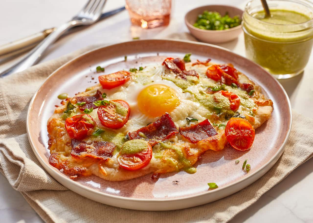 A cooked flatbread, topped with bacon, cheese, cheery tomato halves and an egg, sits on a wide plate, on top of a pale cloth napkin. A glass jar of green sauce sits behind the plate. 
