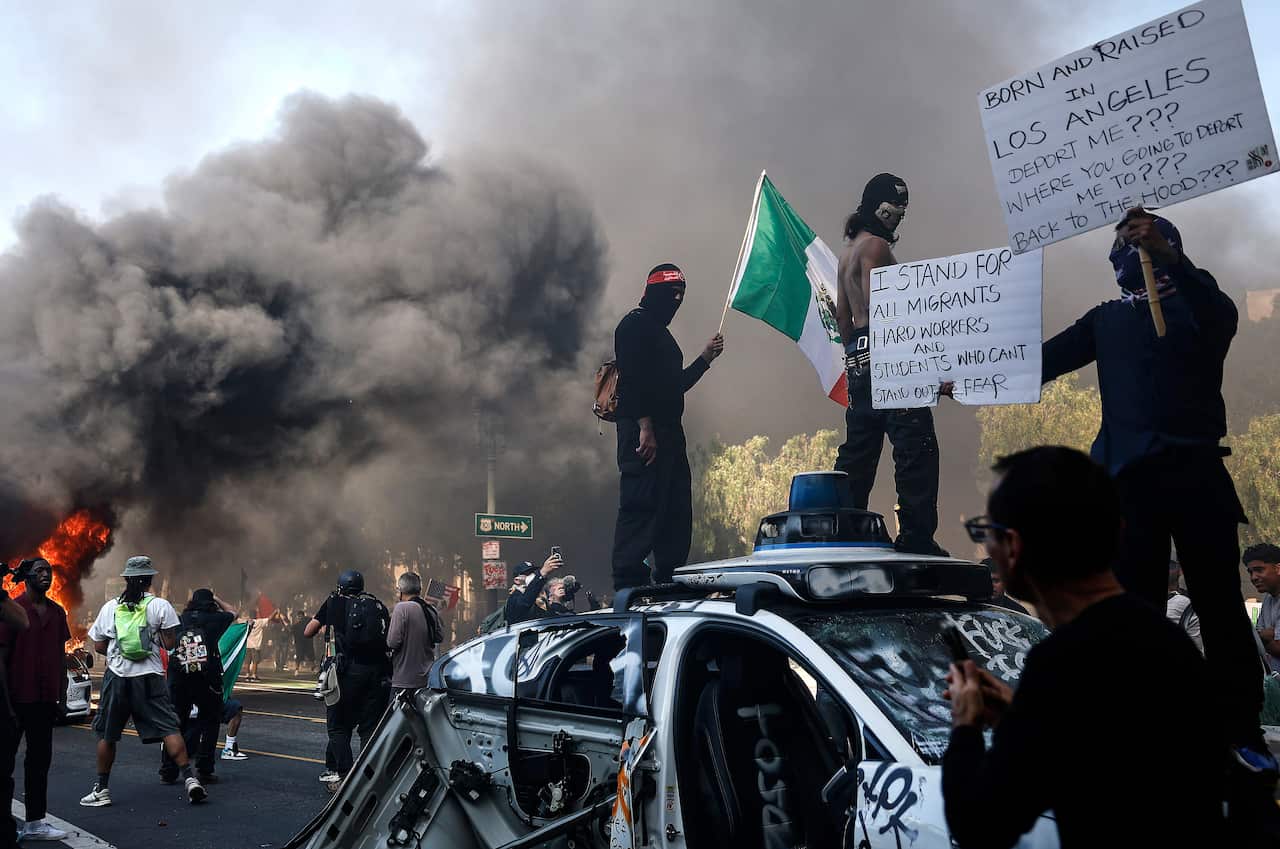 Protesters standing on the roof of a car that has been destroyed, with a cloud of smoke in the distance.