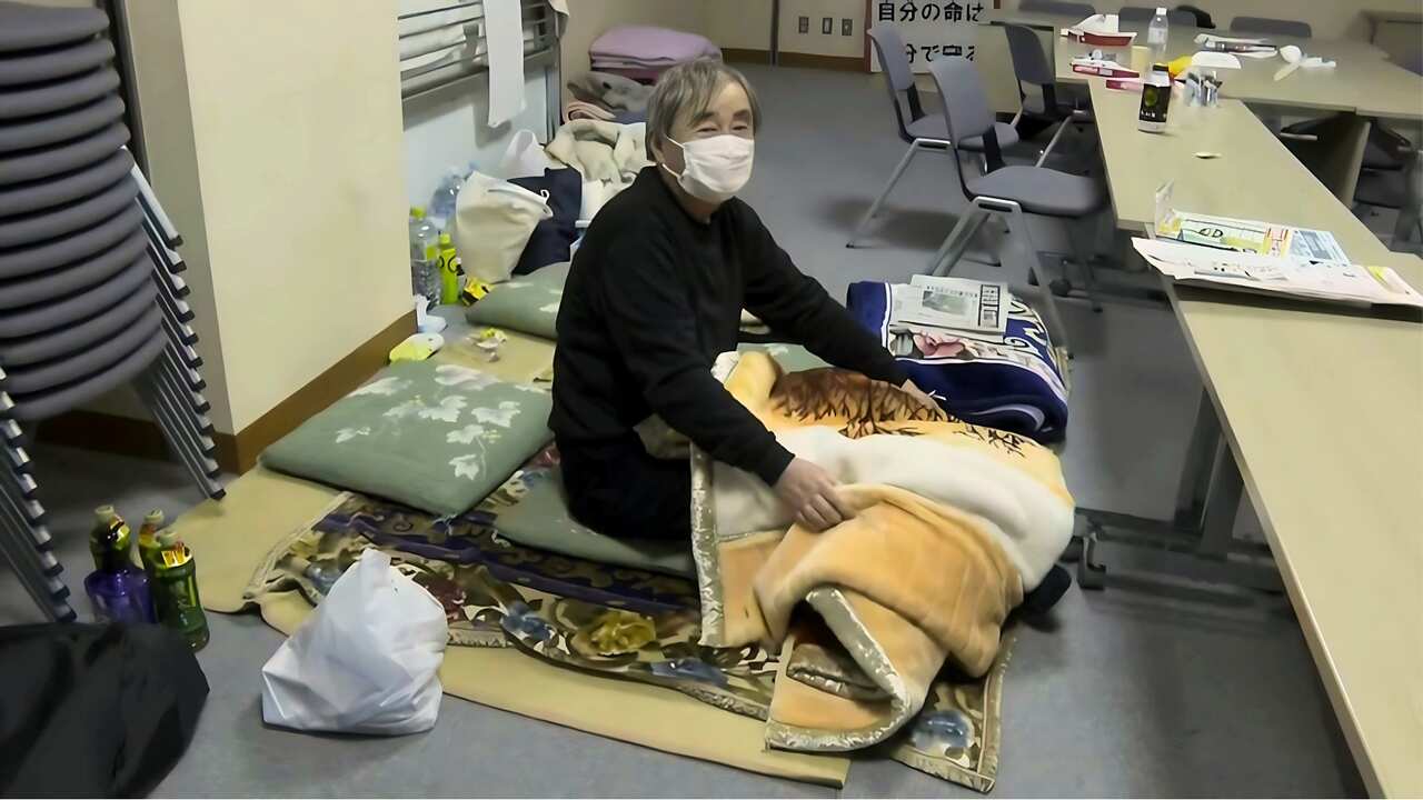 An elderly man in a black sweater and face mask sits on a makeshift bed on the floor. 