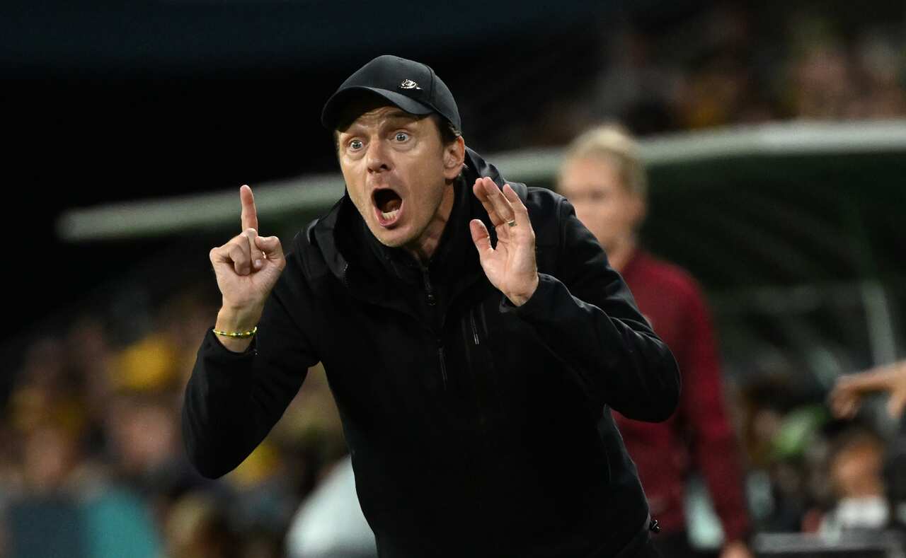 Man in black cap and jacket yells from field sidelines with open mouth, one hand in an open fist and other hand with palm open.
