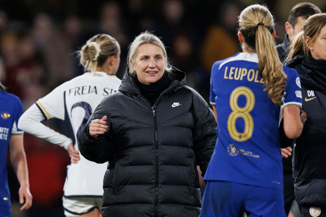 Woman with blonde hair and black puffer jacket speaks to woman in football uniform on field.