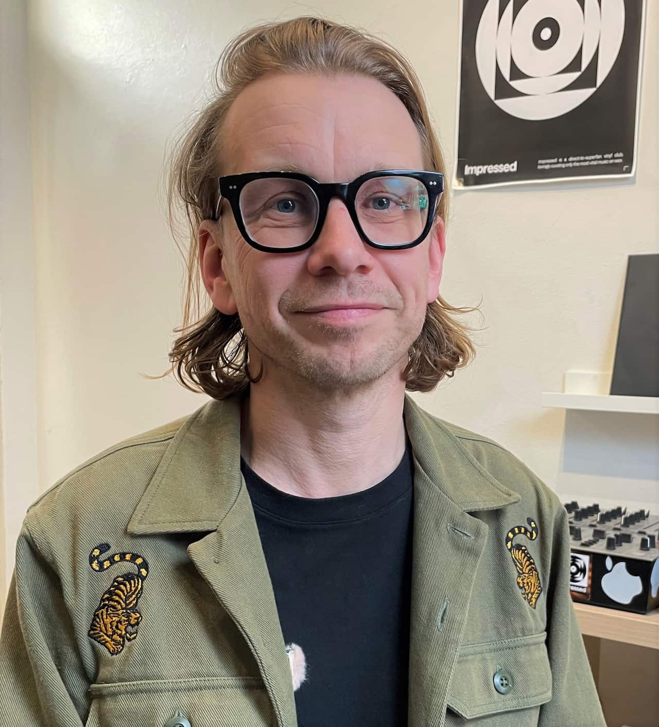 A man wearing black rimmed glasses and a green army jacket sits in an office.