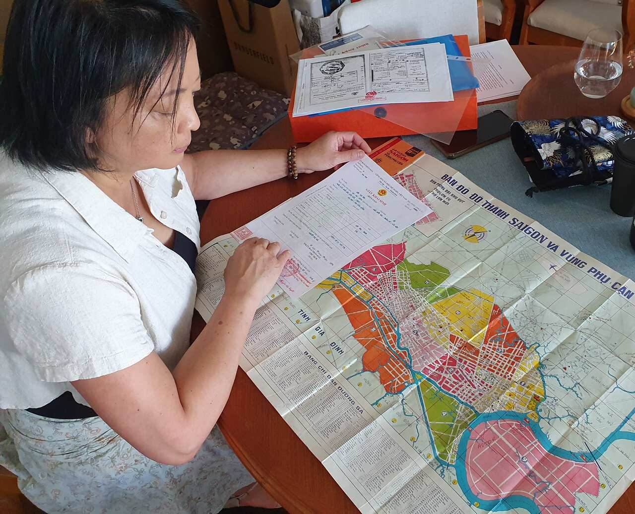 A middle-aged woman with black hair sits at a table looking at an old map of a city.
