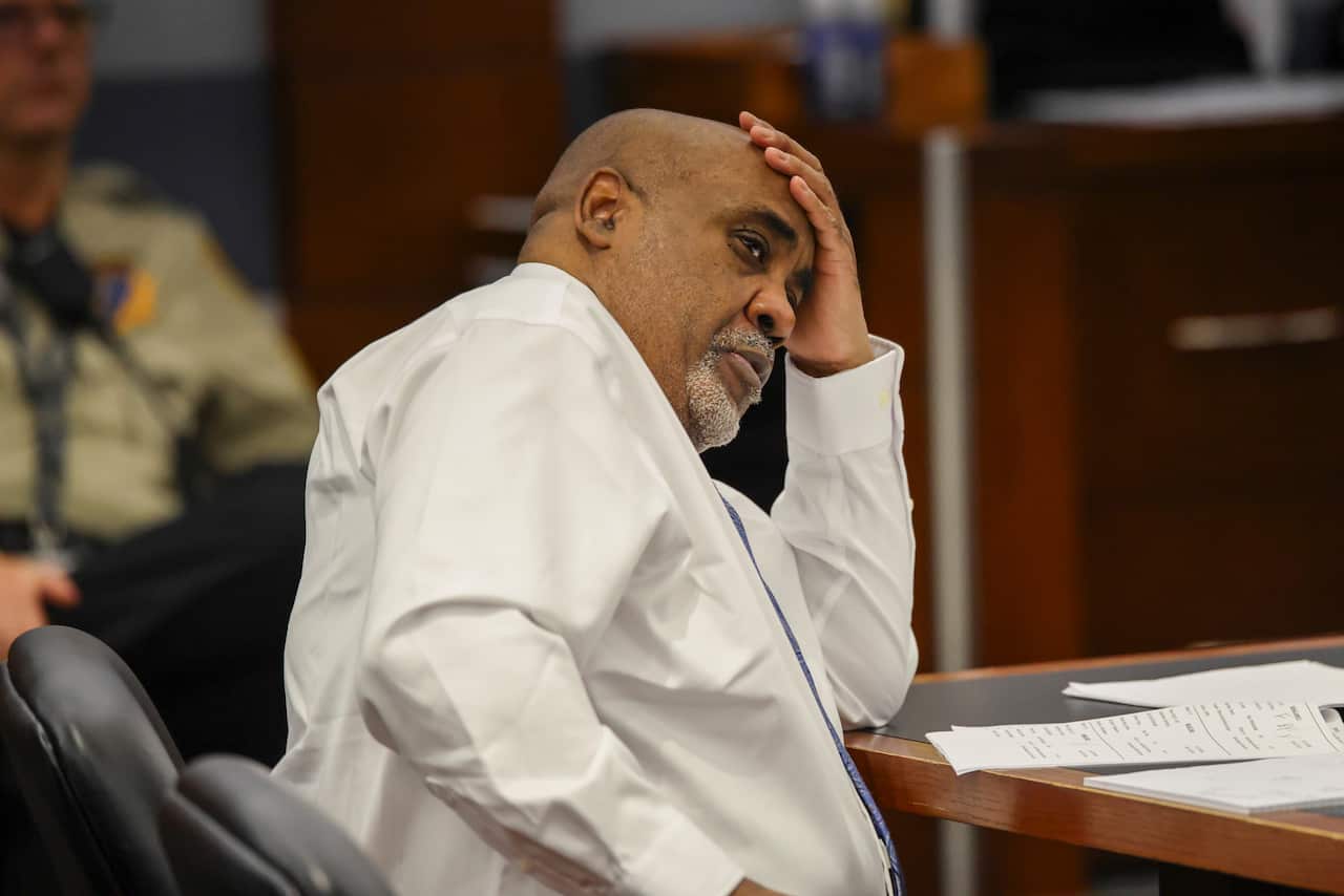 A man wearing a white shirt and a blue tie sits with his hand on his forehead, with his hand resting on the table before him, inside a court.