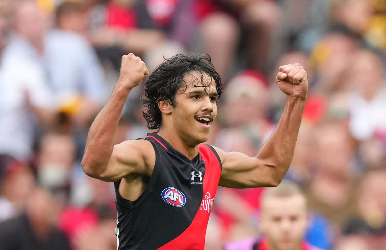 Wearing the black and red jersey of the HAWKS BOMBERS, Alwyn raises his fists and grins in celebration, as an out of focus crowd watches on
