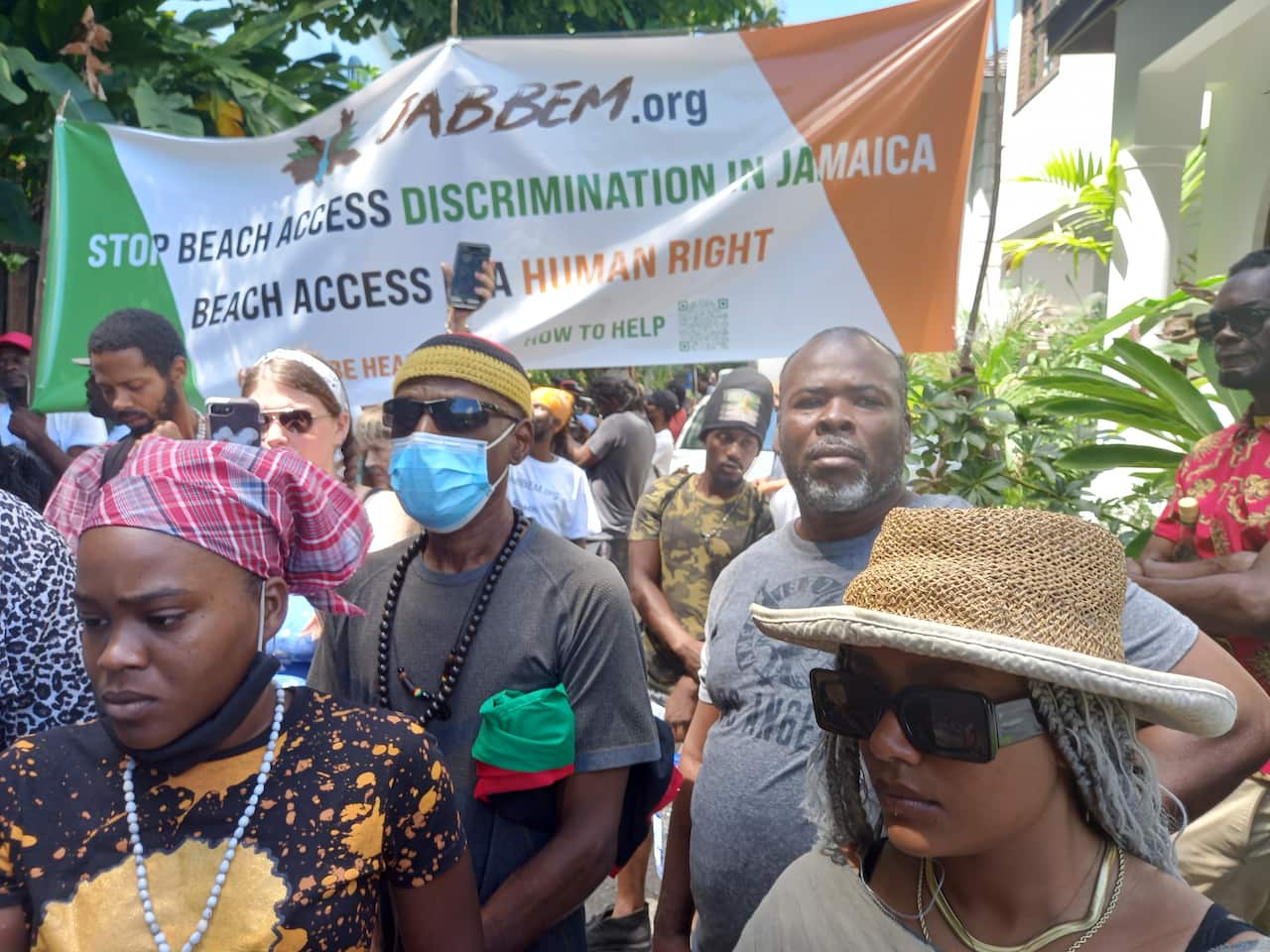 People in hats, masks and sunglasses. There is a large banner behind them that reads in part: Stop beach access discrimination in Jamaica
