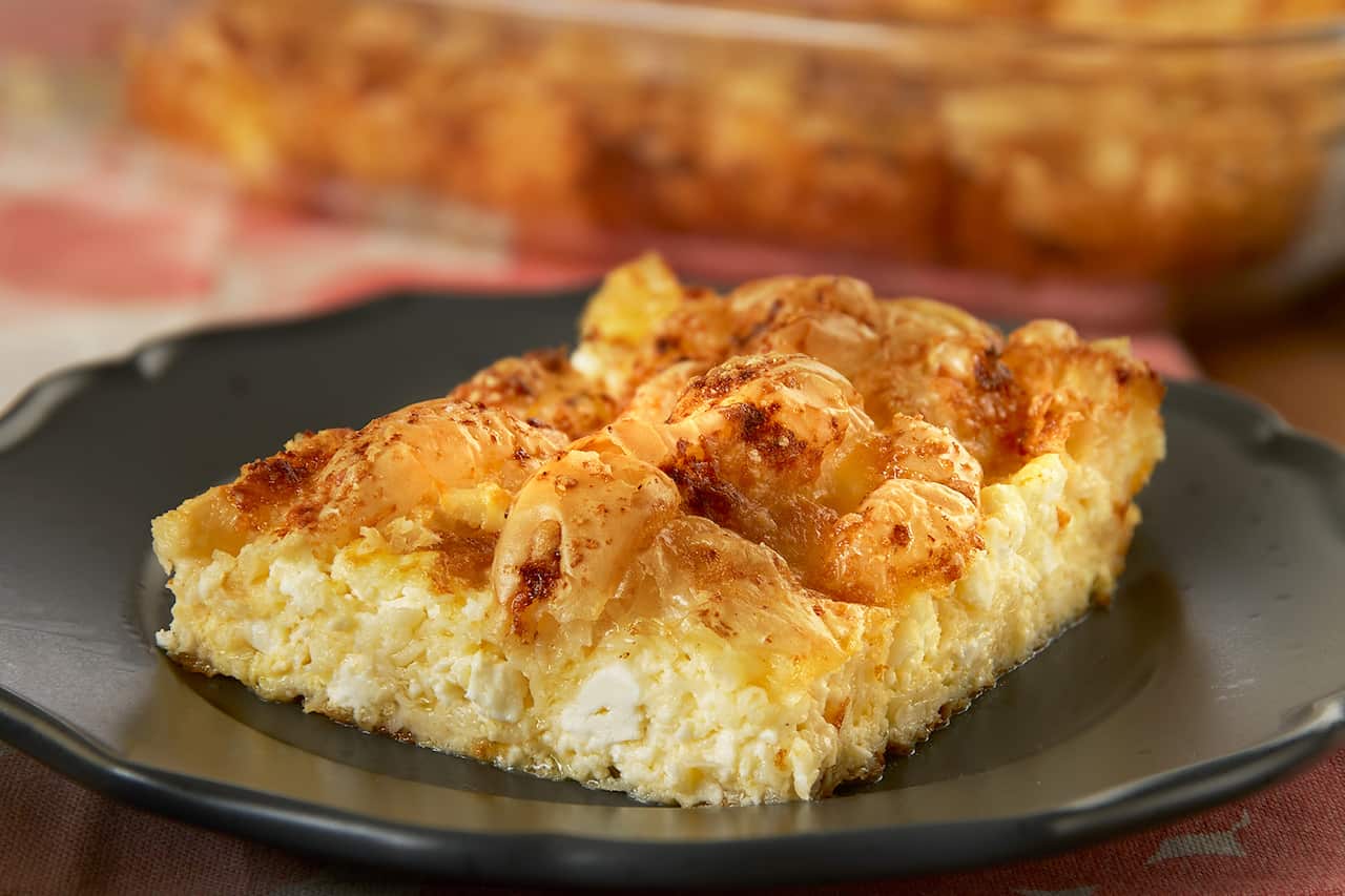 Square piece of pie with soft feta base and slightly scrunched golden-brown filo top sits on dark brown plate. 