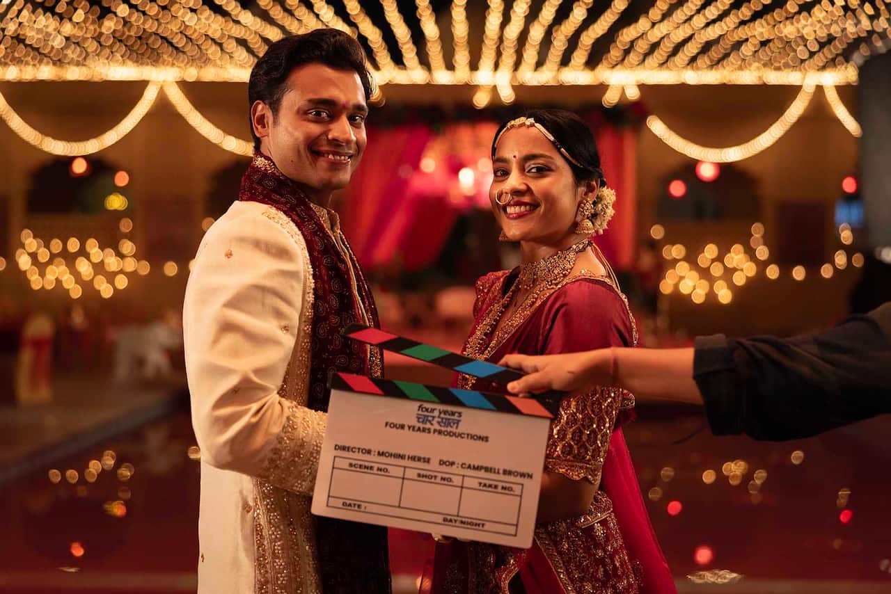 A smiling man and woman in vibrant outfits stand in a glittering room. A film clapper board is held in front of them. 