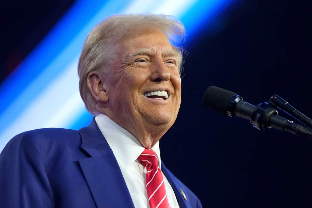 Donald Trump, in a blue suit and red and white striped tie, smiles as he speaks into a microphone.