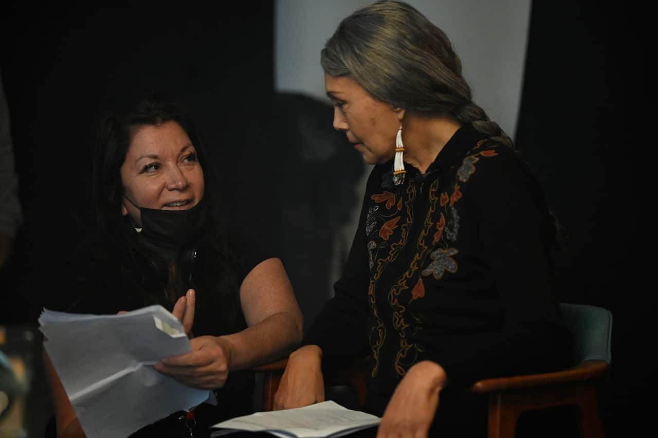Two woman look at each other. One holds several sheets of paper and gestures with her other hand. 