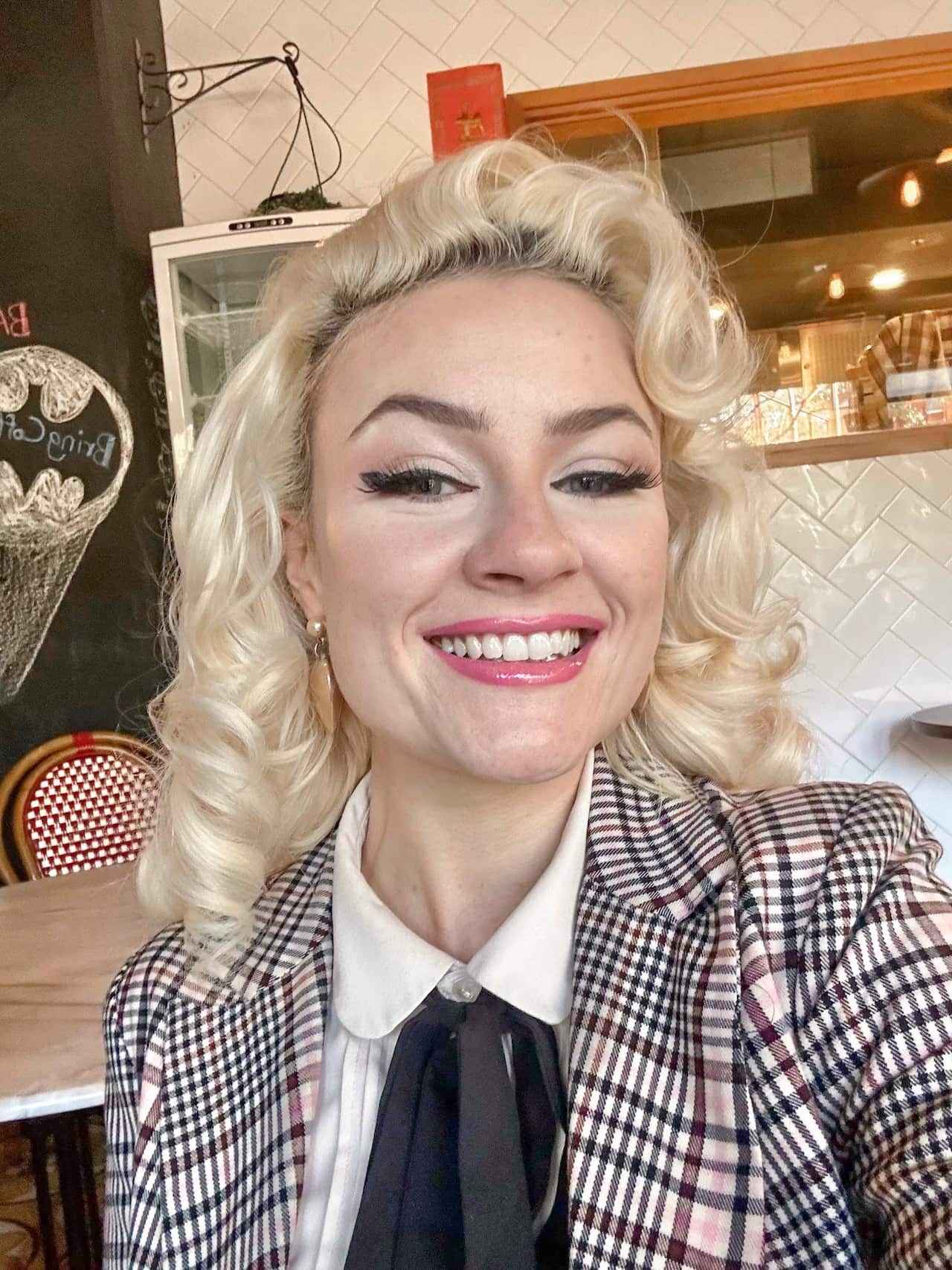 A selfie of a young woman with bleached, 1950s-style hair smiling while wearing a white shirt and tailored checked jacket.