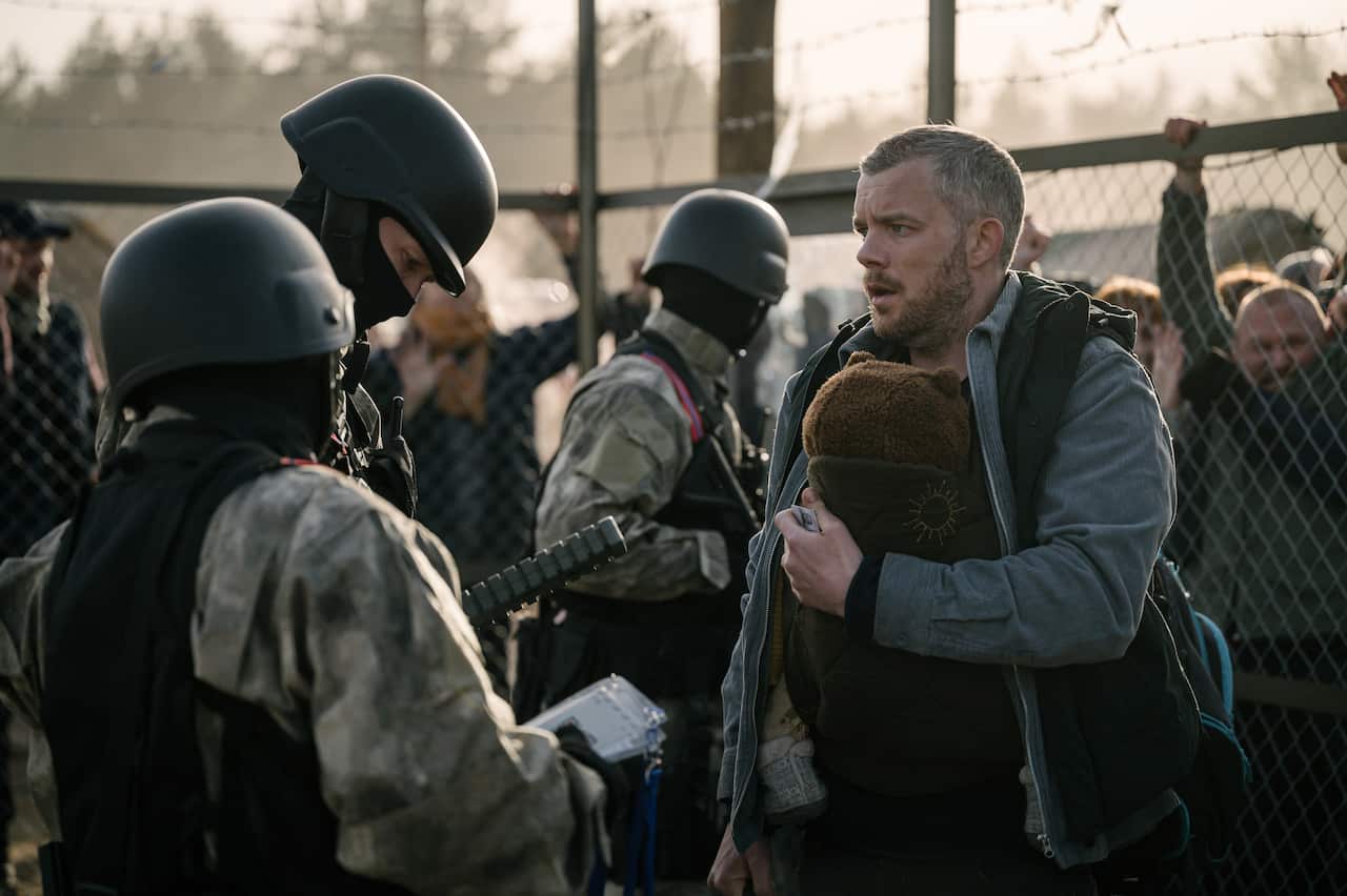 A man stands in front of a chain fence, talking to three people in soldier's uniforms. 