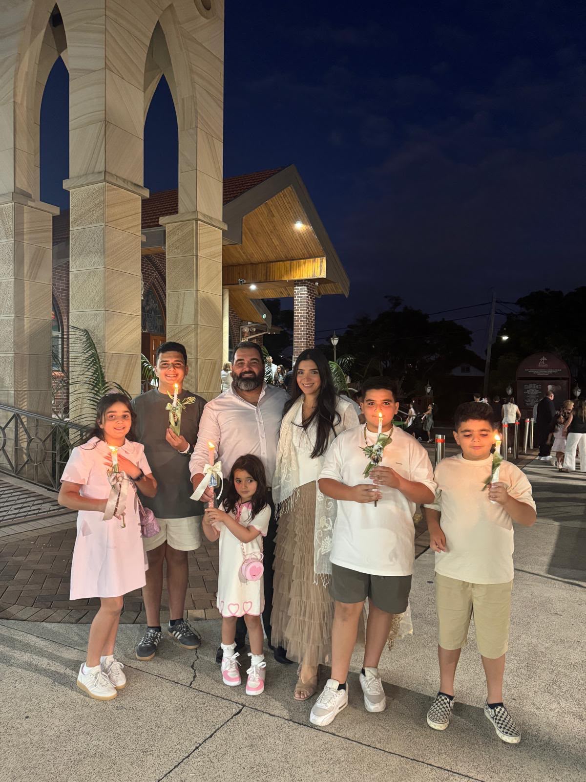 A couple with their five children, holding candles, stands next to Victorian-style pillars outside at night.