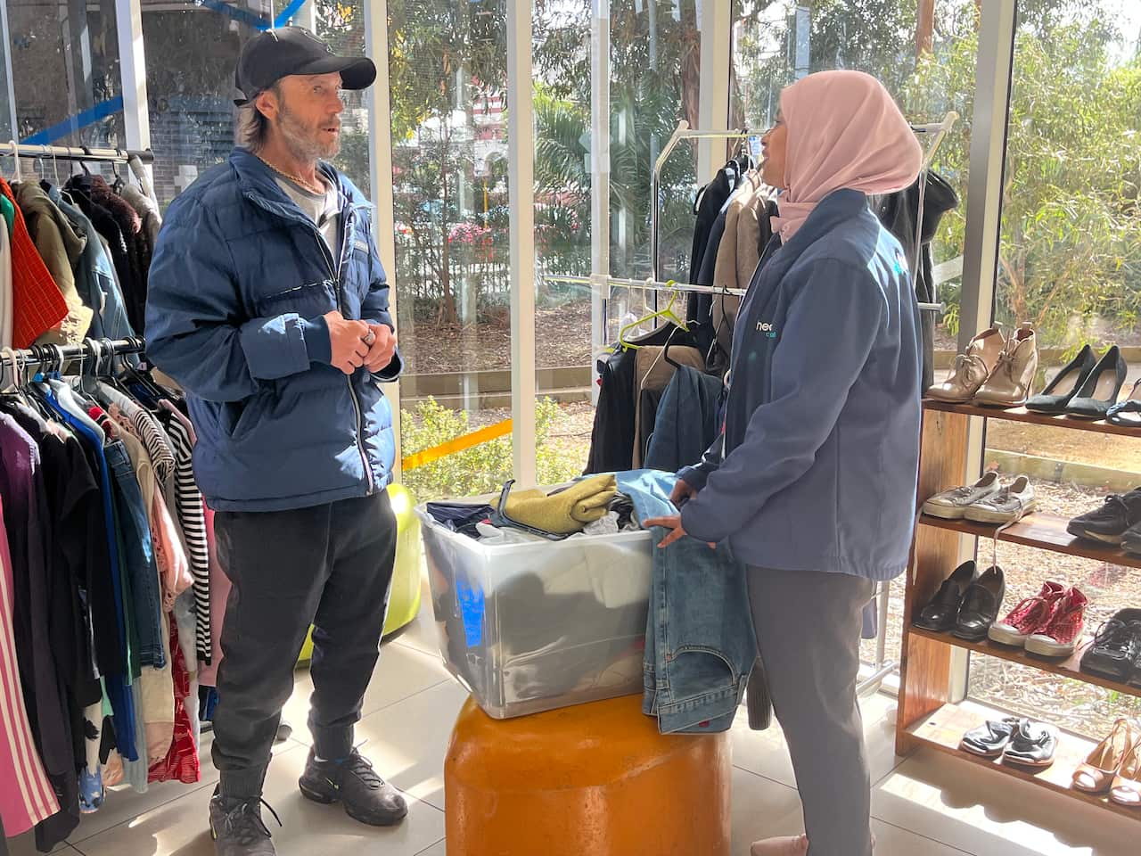 A man talking to a woman. A rack of clothes is behind the man and a rack of shoes is behind the woman.