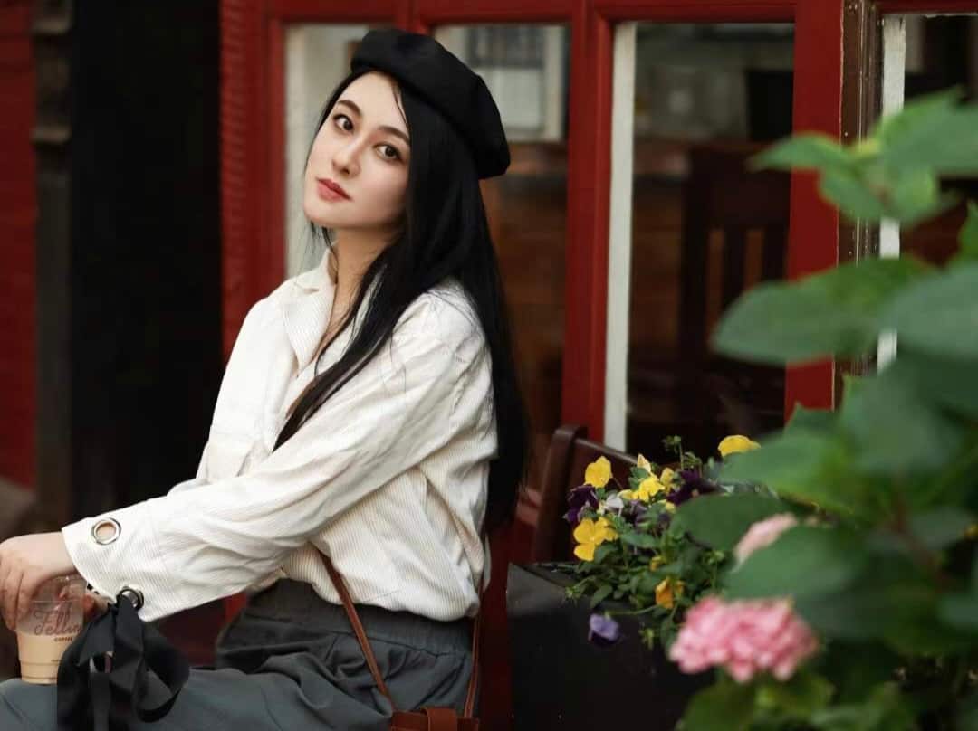 A young woman wearing a black beret and a white button-down shirt sits outdoors at a cafe table beside a planter of colourful flowers.