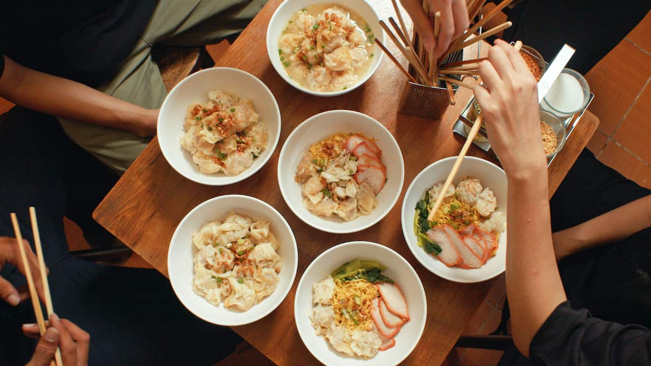 Seen from overhead, a table holds six bowls of food. Hands holding or reaching for chopsticks reach in from the edges of the image.