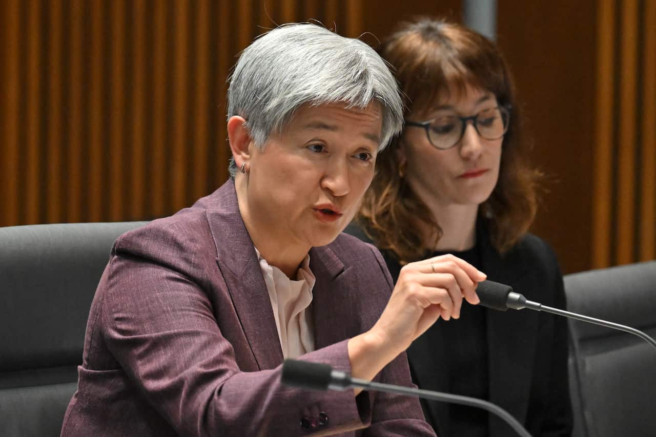 A woman with grey hair speaks into a microphone in a committee room. There's another woman wearing black sitting next to her.