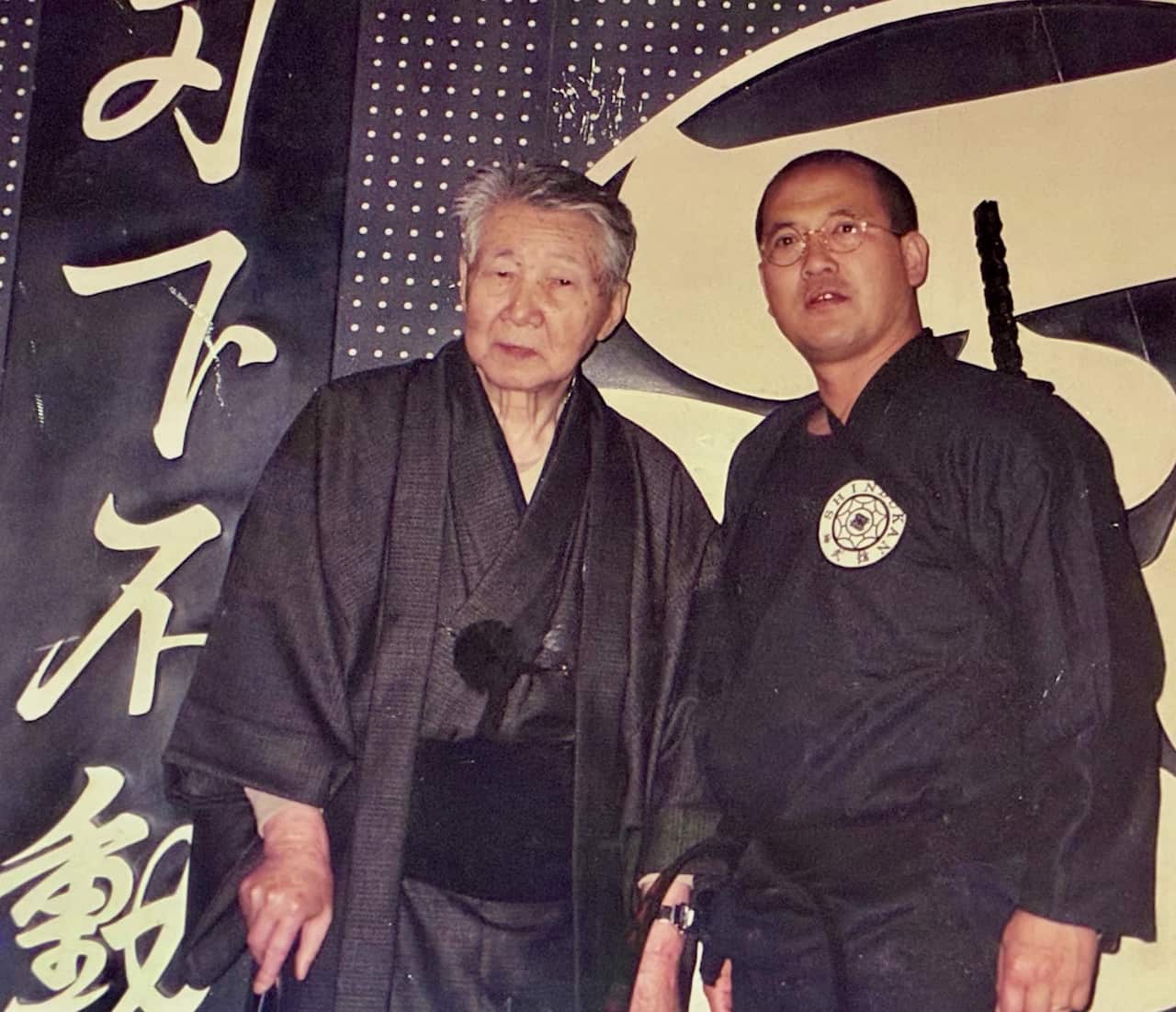 Two men wearing black karate clothing stand facing camera.