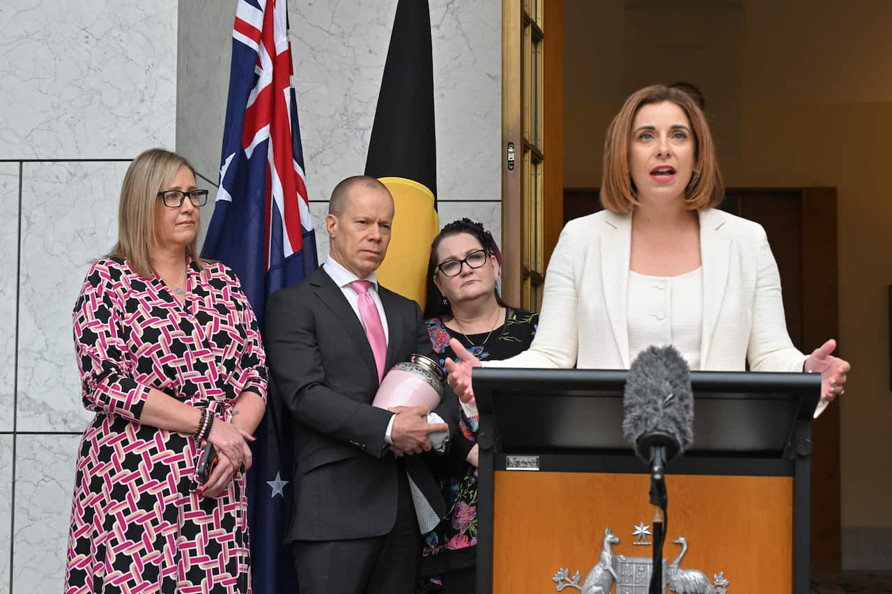 A woman in a white blazer is speaking at a lectern with two women and a man, and flags behind her.