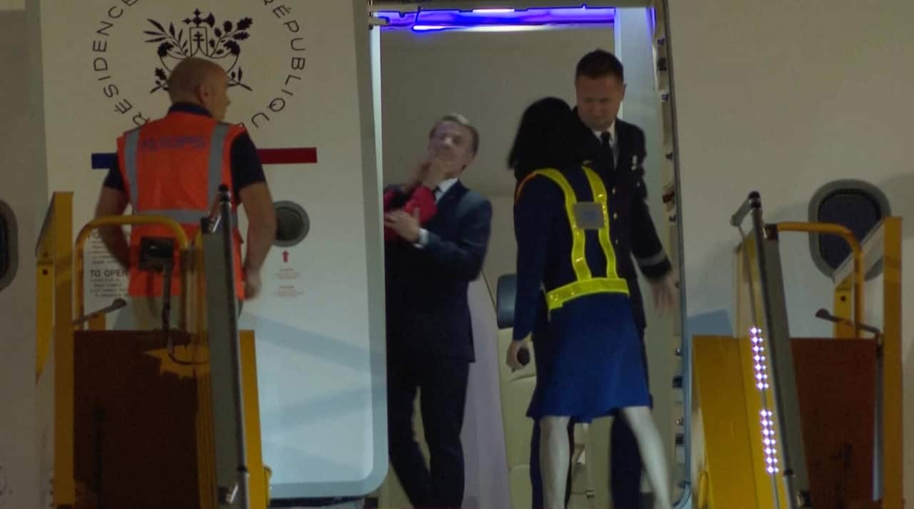 A man in a black suit is being pushed by someone with their hands on his face as he stands at the entrance of an aeroplane. There's a man and a woman in hi vis jackets and a man in a suit around them.