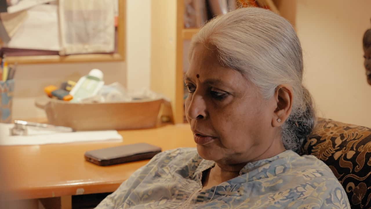 An Indian woman with grey hair in a bun sits and looks ahead with a serious expression on her face.