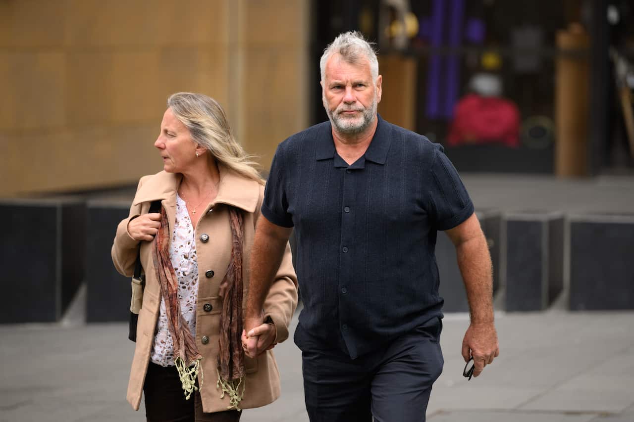 Two people walk hand in hand outside a court building, with one wearing a beige coat and the other a dark short-sleeved shirt.