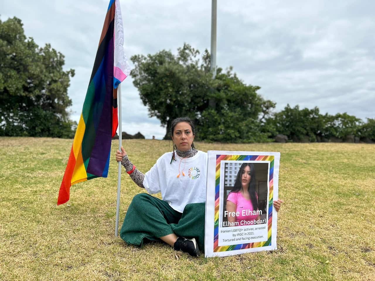 A woman sits on the grass carrying a poster and a pride flag.